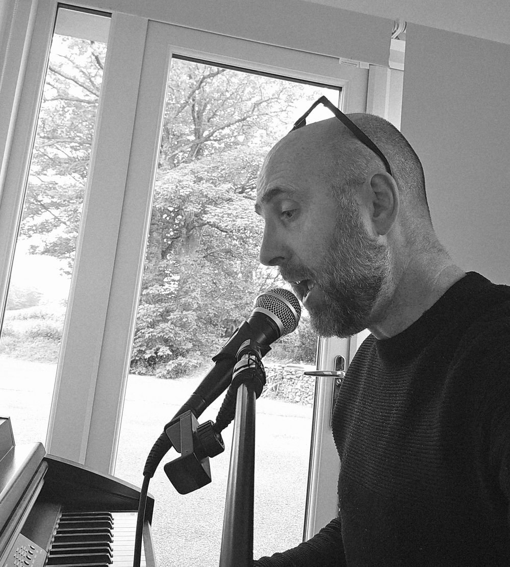 Andy Howie singing into a microphone at the piano by a window